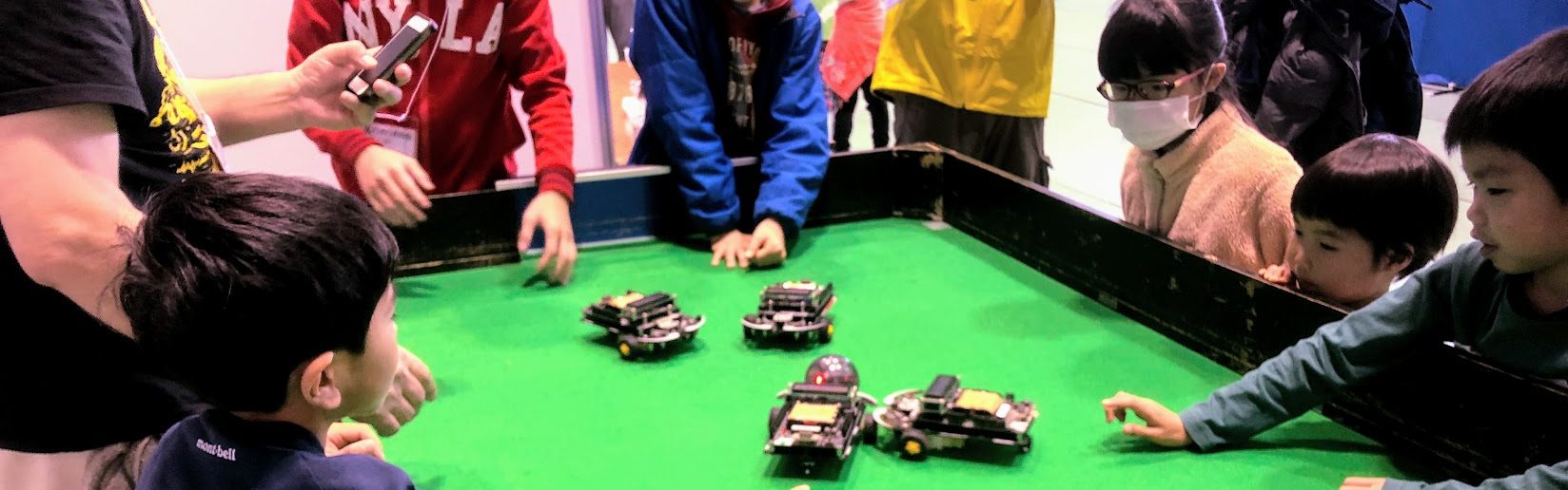 ロボットの競技台を囲み、動きを見守る子どもたち。過去の道場での活動写真。